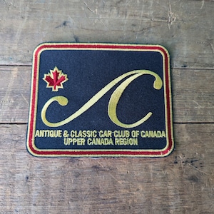 May include: A black embroidered patch with a gold cursive "C" and the text "Antique & Classic Car Club of Canada Upper Canada Region". A red maple leaf is in the top left corner.
