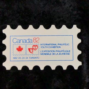 Könnte beinhalten: Eine weiße Gedenkmarke mit blauem und rotem Rand. Die Marke zeigt ein rotes Ahornblatt, ein rotes Herz und den Text "Canada 82" in Blau. Der Text "International Philatelic Youth Exhibition" und "L'Exposition Philatélique Mondiale de la Jeunesse" ist ebenfalls in Blau enthalten. Das Datum "1982 05.20-24 TORONTO" ist unten in Blau gedruckt.