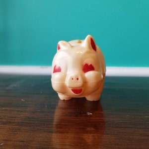 May include: A white plastic piggy bank with red heart-shaped eyes and a red smiling mouth.