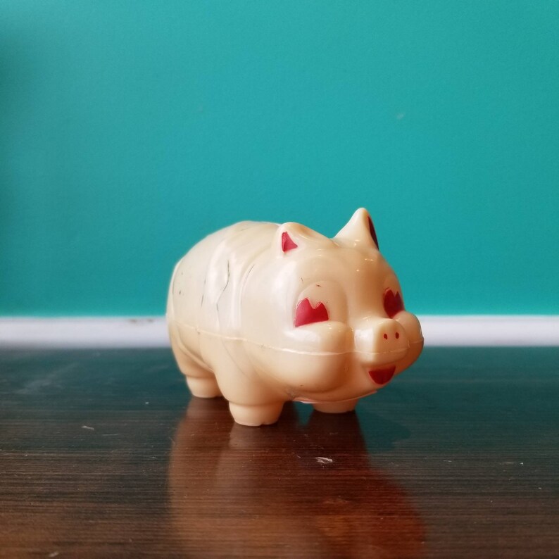 May include: A white plastic piggy bank with red painted eyes and a smiling mouth. The piggy bank is sitting on a wooden surface.