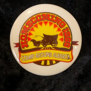 Könnte beinhalten: Ein weißer Knopf mit einem roten und gelben Design. Der Knopf zeigt einen von Pferden gezogenen Wagen mit dem Text "Homecoming 1980" und "Stamp Around Alberta".