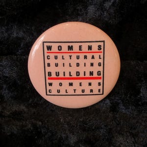 Könnte beinhalten: Ein rosa Knopf mit den Worten "WOMENS CULTURAL BUILDING BUILDING WOMENS CULTURE" in schwarzer und roter Schrift.