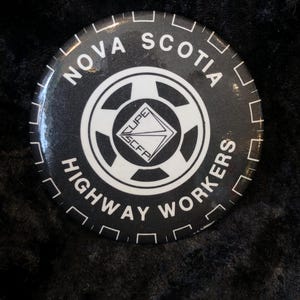 Könnte beinhalten: Ein schwarz-weißer Knopf mit dem Text "Nova Scotia Highway Workers" und einem Logo mit einem Diamanten, in dem die Buchstaben "CUPE" stehen.