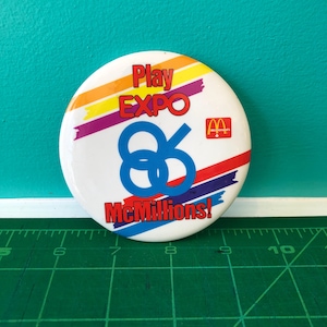 Könnte beinhalten: Ein runder, weißer Button mit dem Text "Play EXPO 86 McMillions!" in Rot, Blau und Gelb. Das McDonald's-Logo ist zu sehen. Das Design ist retro.