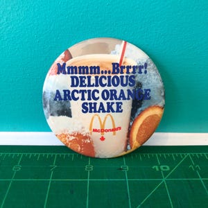 Könnte beinhalten: Ein runder Button mit Werbung für einen McDonald's Arctic Orange Shake. Der Button zeigt den Text "Mmmm...Brrrr! DELICIOUS ARCTIC ORANGE SHAKE" in Blau und Weiß. Eine Orangenscheibe und ein roter Strohhalm sind ebenfalls abgebildet.