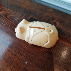 May include: A white ceramic piggy bank with a slot on the back. The piggy bank has a stylised design on its back and red painted eyes.