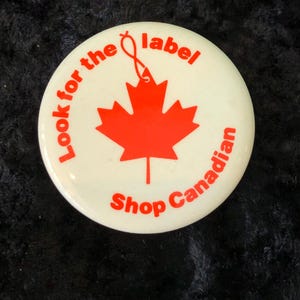 Könnte beinhalten: Ein weißer Knopf mit rotem Text und einem roten Ahornblatt. Der Text lautet "Look for the label Shop Canadian".
