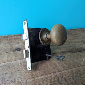 May include: Antique brass door knob and lock set with a key. The lock has a white painted plate and three visible locking positions. The knob is round and has a slightly textured surface.