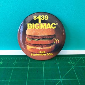 Könnte beinhalten: Ein runder Button mit einem Big Mac Burger und dem Text "$1.39 BIG MAC" in Gelb und Weiß. Das McDonald's Logo und der Text "Bis zum 30. September" sind ebenfalls vorhanden. Der Button befindet sich vor einem türkisfarbenen Hintergrund.
