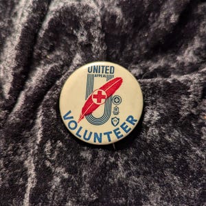 May include: A white button with a red cross and a blue feather inside a blue "U" with the text "UNITED APPEAL VOLUNTEER" around the outside.