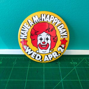 Könnte beinhalten: Ein runder, gelber Button mit Ronald McDonald mit roten Haaren und rotem Lächeln. Der Button zeigt "Have a McHappy Day! Wed. Apr. 2" in einem kreisförmigen Design. Zwei rote Sterne sind an den Seiten.