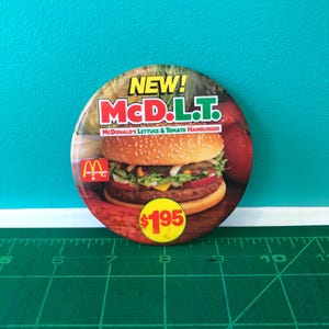 Könnte beinhalten: Ein runder Button mit einem McDonald's McDLT-Hamburger. Der Button trägt oben den Text "NEW! McDLT", das McDonald's-Logo und den Preis 1,95 $. Das Bild zeigt eine Nahaufnahme des Burgers mit Salat und Tomate.