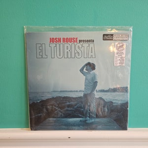 Op de afbeelding: Een vinyl platenhoes met een man in een wit overhemd en hoed die op een houten pier staat en uitkijkt over een waterpartij. De albumtitel is "EL TURISTA" en de artiest is "JOSH ROUSE presenta".