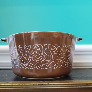 May include: A brown ceramic casserole dish with a white floral design. The dish has two handles and a rounded shape.