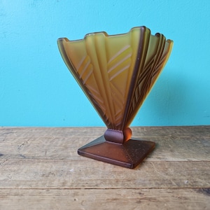 Könnte beinhalten: Eine Vase im Art-Déco-Stil in Bernstein- und Brauntönen. Die fächerförmige Oberseite weist geätzte geometrische Muster auf. Die Vase steht auf einem quadratischen Sockel und hat einen runden Akzent. Der Hintergrund ist türkis.