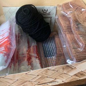 May include: A craft kit for rug hooking with a variety of materials including black yarn, orange and white yarn, brown yarn, and a canvas grid.