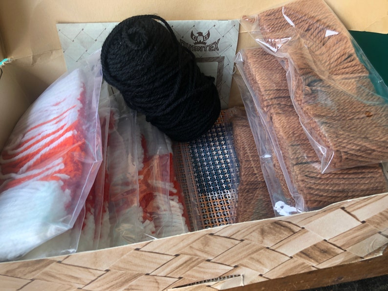 May include: A craft kit for rug hooking with a variety of materials including black yarn, orange and white yarn, brown yarn, and a canvas grid.