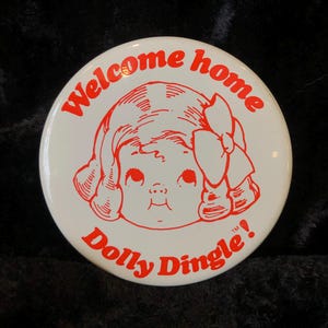 Könnte beinhalten: Weißer Knopf mit rotem Text und dem Gesicht eines Zeichentrickmädchens. Der Text lautet "Welcome home Dolly Dingle!"