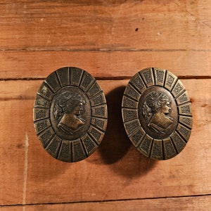 Może przedstawiać: Dwa antyczne mosiężne gałki do szafek z wypukłym wzorem profilu kobiety w kółku otoczonym ozdobną ramką.