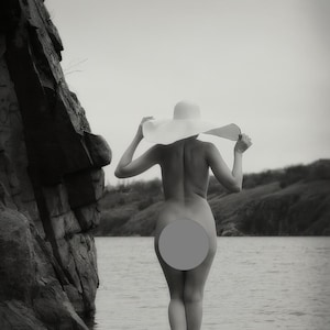May include: A woman wearing a large white sun hat stands on a rock in a lake. The water is calm and the background is a rocky cliff.