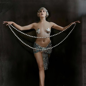 Puede incluir: Una mujer con una falda de cuentas y un collar de perlas posa para una foto. Lleva un tocado decorativo y tiene un fondo oscuro.