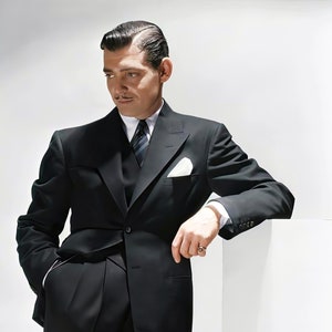 Puede incluir: Un hombre con un traje negro, un pañuelo de bolsillo blanco y una corbata oscura. Está de pie con una mano en el bolsillo y la otra apoyada en una superficie blanca.