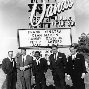 Puede incluir: Foto en blanco y negro de cinco hombres con traje de pie frente al Sands Hotel en Las Vegas. El letrero sobre ellos dice "Sands A Place In The Sun". Un letrero debajo del letrero enumera los nombres de los artistas: Frank Sinatra, Dean Martin, Sammy Davis Jr., Peter Lawford y Joey Bishop.