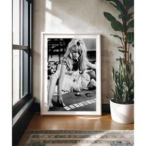 May include: Black and white framed print featuring a woman playing cards. The woman is wearing a white dress and smoking a cigarette. The print is displayed in a room with a window, a plant, and a rug.