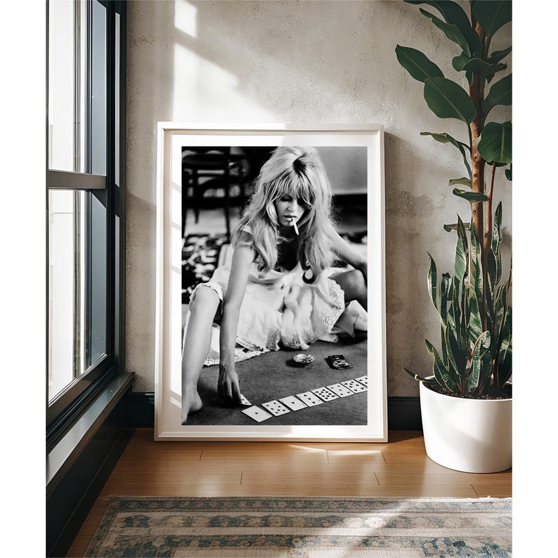 May include: Black and white framed print featuring a woman playing cards. The woman is wearing a white dress and smoking a cigarette. The print is displayed in a room with a window, a plant, and a rug.