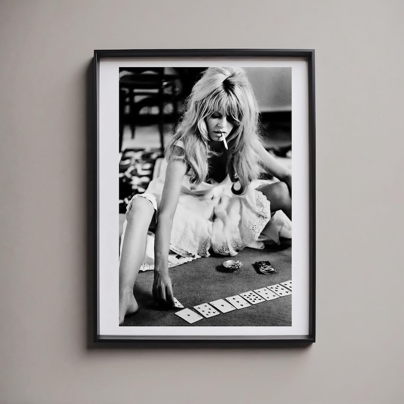 May include: Black and white framed print featuring a woman with long hair, smoking a cigarette, playing dominoes. The image is a vintage photograph, with a black frame. The woman is wearing a white dress and is sitting on the floor.