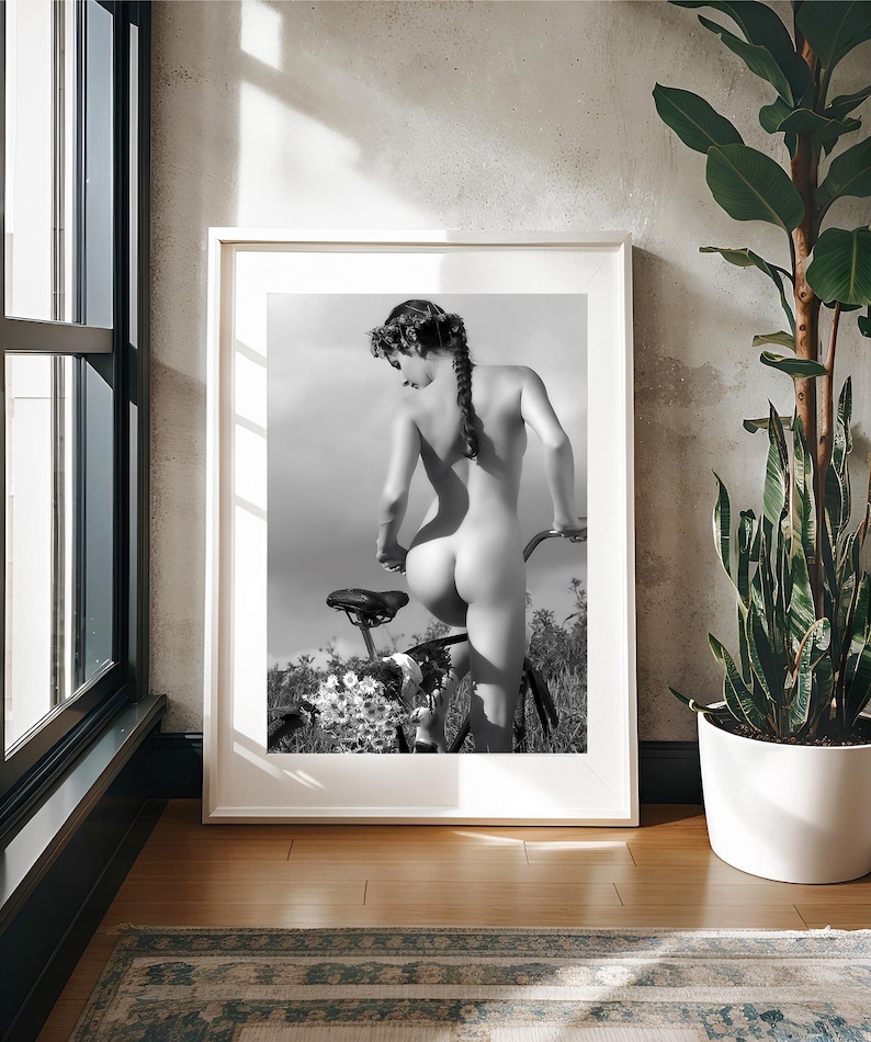 May include: A black and white photograph of a person standing behind a bicycle. The person is nude and has their back to the camera. The bicycle is in the background and is partially obscured by flowers.