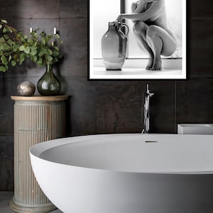 May include: A white freestanding bathtub with a silver tiled base in a bathroom with dark grey tiled walls. A framed black and white photograph of a woman sitting on a ledge with a large ceramic vase is hanging on the wall.