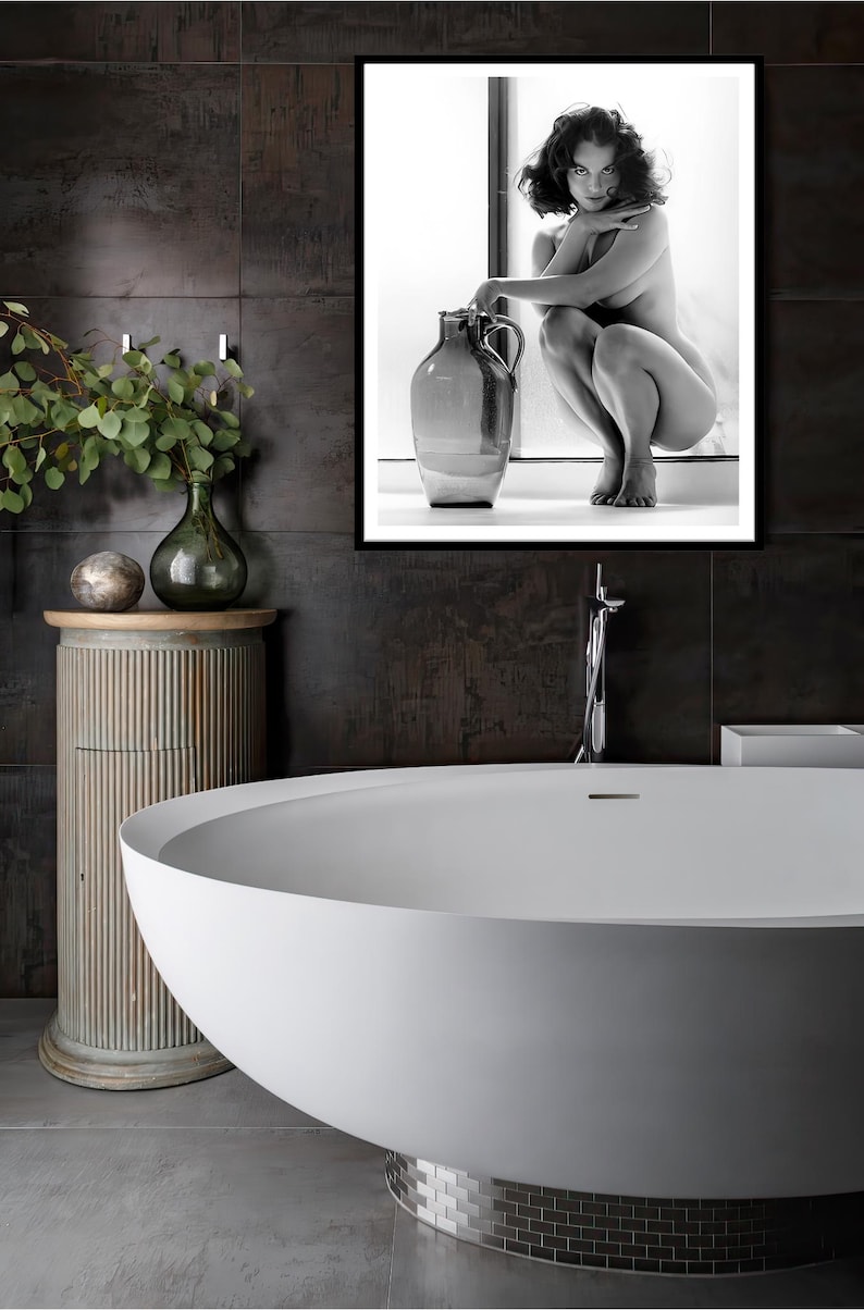 May include: A white freestanding bathtub with a silver tiled base in a bathroom with dark grey tiled walls. A framed black and white photograph of a woman sitting on a ledge with a large ceramic vase is hanging on the wall.