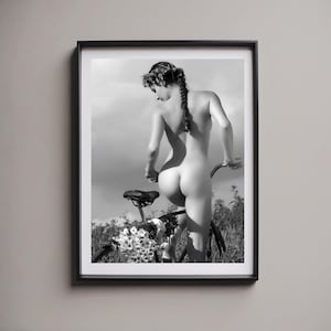 May include: A black and white photograph of a person standing on a bicycle with a floral arrangement on the handlebars. The person is nude and has a flower crown in their hair.