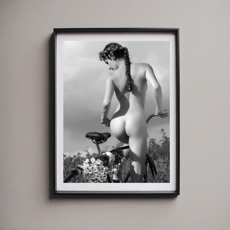May include: A black and white photograph of a person standing on a bicycle with a floral arrangement on the handlebars. The person is nude and has a flower crown in their hair.