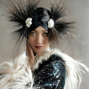 May include: A woman wearing a black turban-style headpiece adorned with black feathers and pearl-centered floral accents. She is wearing a sequined jacket with white feather trim.