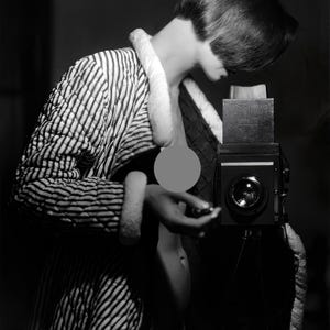 Puede incluir: Una fotografía en blanco y negro de una persona que lleva una bata a rayas y sostiene una cámara vintage. La cámara está en un trípode y la persona está mirando hacia abajo a la cámara.