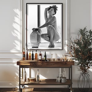 Peut inclure: Une photographie en noir et blanc d'une femme posant avec une grande jarre en c&eacute;ramique. La femme est nue et a les cheveux tir&eacute;s en arri&egrave;re. La photographie est encadr&eacute;e et accroch&eacute;e &agrave; un mur blanc au-dessus d'un chariot de bar en bois avec des bouteilles et des verres.