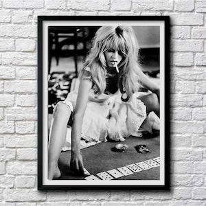 May include: Black and white framed print of a woman with long hair, smoking a cigarette, playing cards. The woman is wearing a white dress and is seated on the floor. A deck of cards is laid out in front of her.