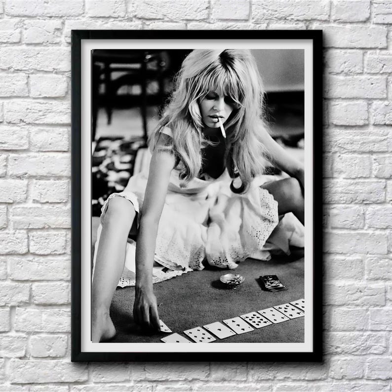 May include: Black and white framed print of a woman with long hair, smoking a cigarette, playing cards. The woman is wearing a white dress and is seated on the floor. A deck of cards is laid out in front of her.