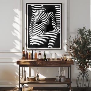 May include: A black and white photograph of a person's torso with light and shadow stripes. The photo is framed and hanging on a white wall above a wooden bar cart with bottles and glasses.