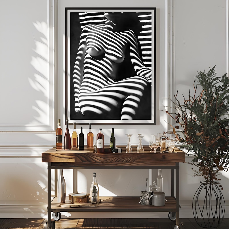 May include: A black and white photograph of a person's torso with light and shadow stripes. The photo is framed and hanging on a white wall above a wooden bar cart with bottles and glasses.