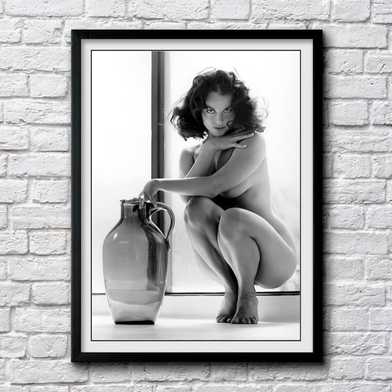 May include: A black and white photograph of a woman sitting with her legs crossed, holding a large ceramic jug. The woman is nude and has her hair pulled back. The background is a white wall with a window.