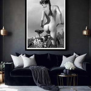 May include: A black and white photograph of a person standing behind a bicycle. The person is nude and has a flower crown in their hair. The bicycle is in the background and is partially obscured by flowers.