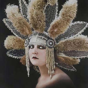 May include: A woman wearing a large, elaborate headdress with feathers and beads. The headdress is white and silver with brown feathers. The woman is wearing heavy eye makeup and a dark red lipstick.
