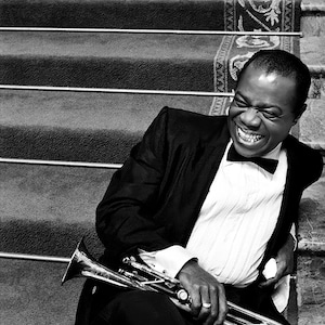 May include: Black and white photograph of a smiling person in a tuxedo, holding a trumpet. The person is seated on a staircase with metal railings. The trumpet is silver and the person is wearing a bow tie.