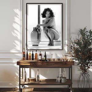 May include: A black and white photograph of a woman sitting on a ledge with a large ceramic jug. The woman is nude and has her hair pulled back. The photograph is framed in a black frame and is hanging on a white wall.