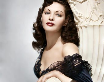 May include: A woman with dark brown hair and red lipstick is wearing a black off-the-shoulder gown with lace detailing. She is sitting in a white ornate chair with a sculpted figure on the back.