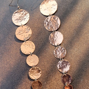 May include: A pair of long, hammered copper earrings with multiple circular discs hanging from the top. The earrings are a warm, reddish-brown color.