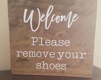 May include: A wooden sign with the text "Welcome Please remove your shoes" in white lettering on a brown background.